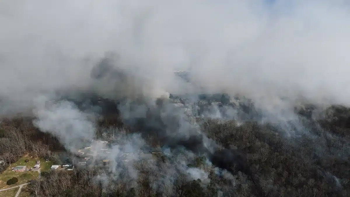 A massive orange wildfire advancing through dry grassland and timber, smoke billowing into a hazy sky, flames consuming vegetation near a distant highway corridor, extreme heat and motion visible