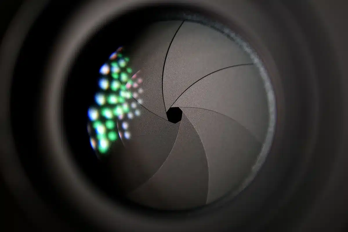 A smartphone camera lens assembly with mechanical aperture blades visible through the lens, adjustable metal rings catching light, precision engineering detail