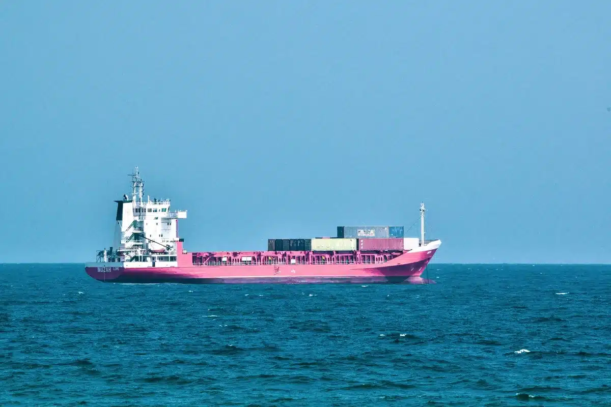 A narrow maritime chokepoint with cargo vessels anchored in shallow turquoise waters, flanked by rocky coastlines, one side arid and tan, the other distant and hazy, showing the confined geography of global trade