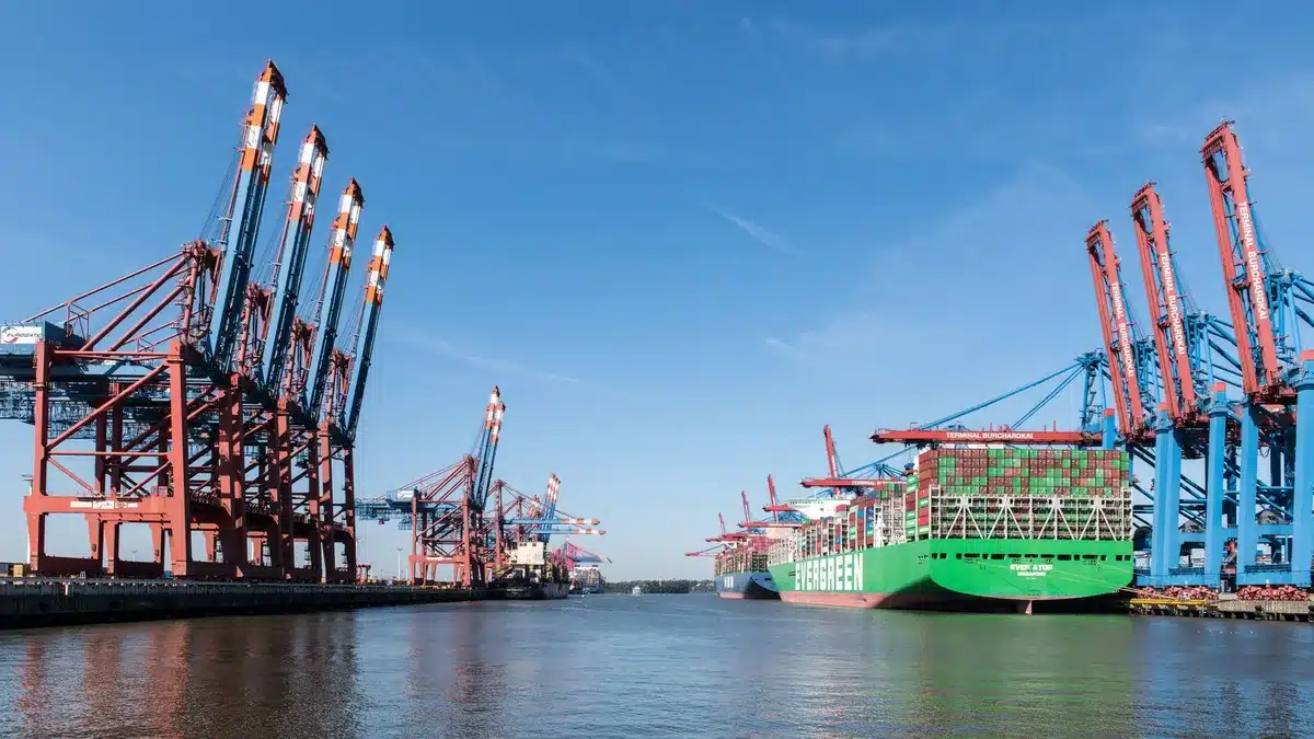 Empty container terminal at dusk with stacked cargo boxes, cranes idle, few workers visible, industrial landscape showing economic stagnation