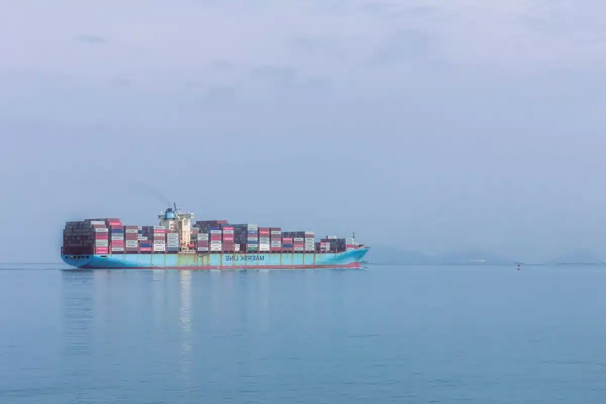 A container ship navigating through open waters, morning sunlight on calm sea, distant coastline visible, maritime commerce resuming