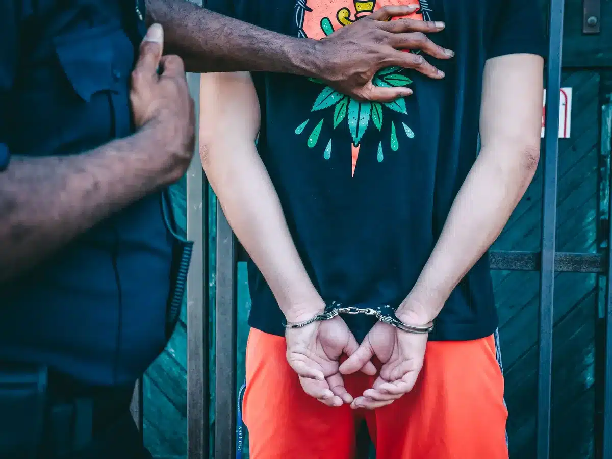 Police station interrogation room with anonymous woman in custody, hands visible on table, officer standing nearby, harsh fluorescent lighting, tense atmosphere