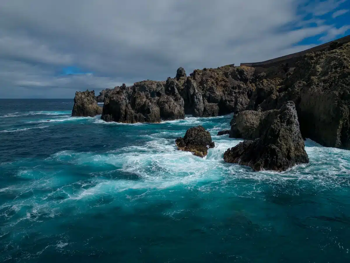 A wide view of a rocky volcanic coastline with crashing waves, steep cliffs rising from the ocean, no people visible, representing the Izu Islands' volcanic geology and seismic setting