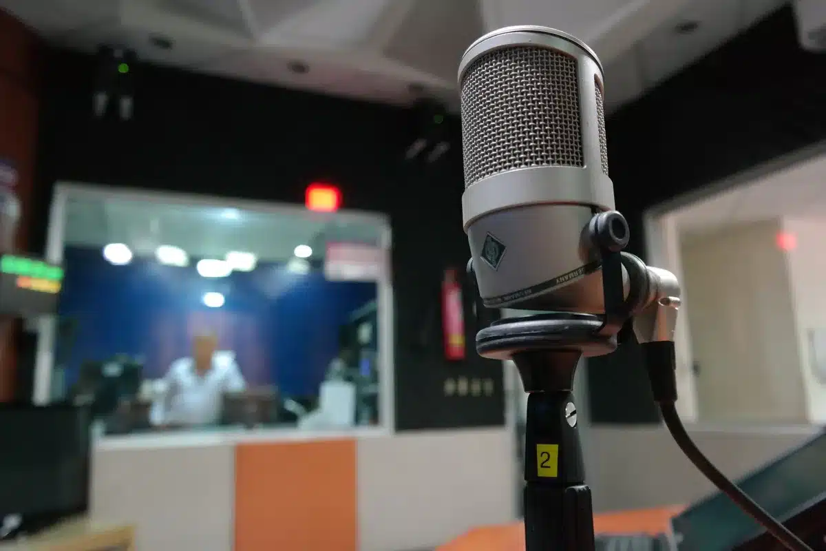 A red-curtained podcast studio with a microphone under stage lighting, recording equipment visible on a desk, minimal decor emphasizing the intimate interview setting