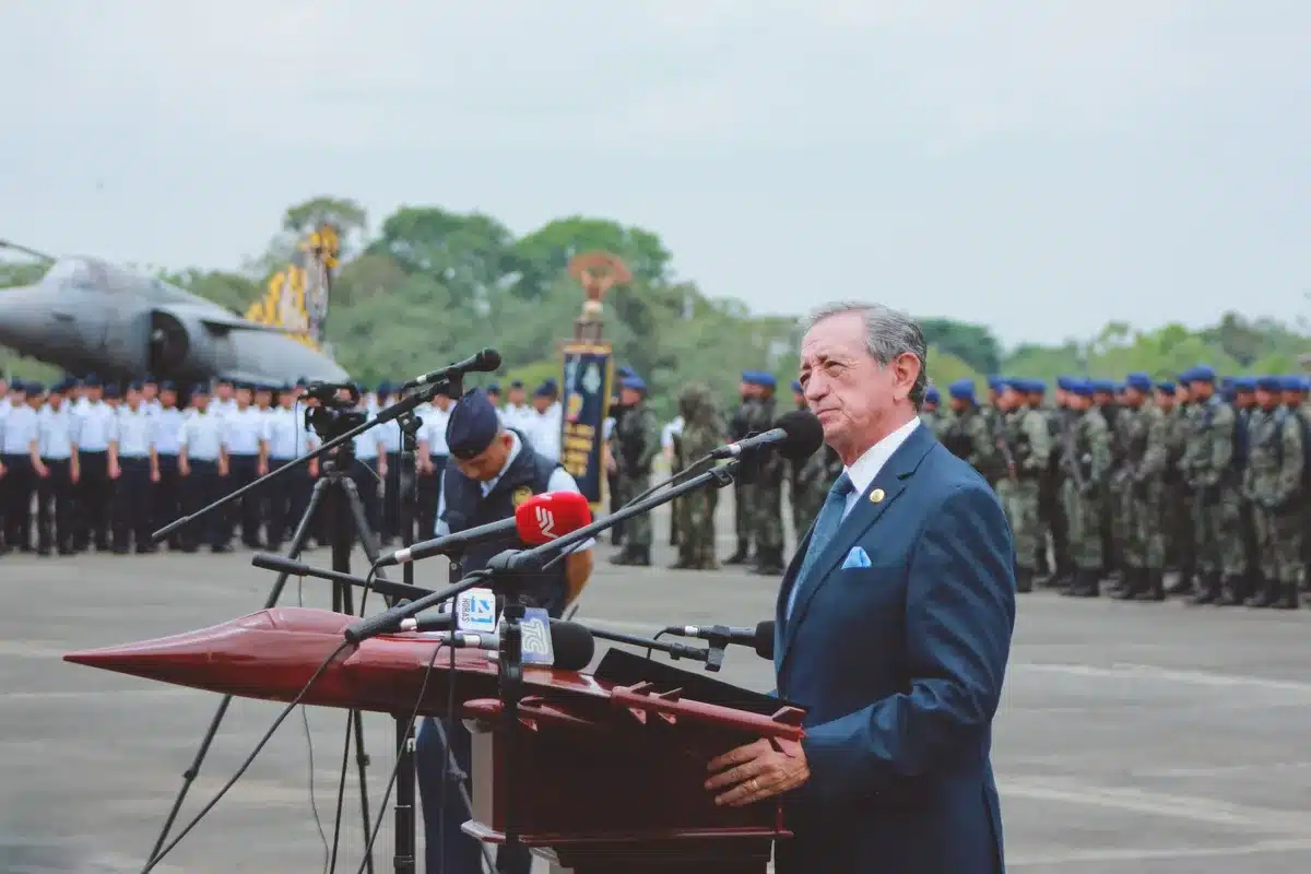 Military personnel standing at attention during an outdoor ceremony, flags visible in background, a podium with microphones in focus