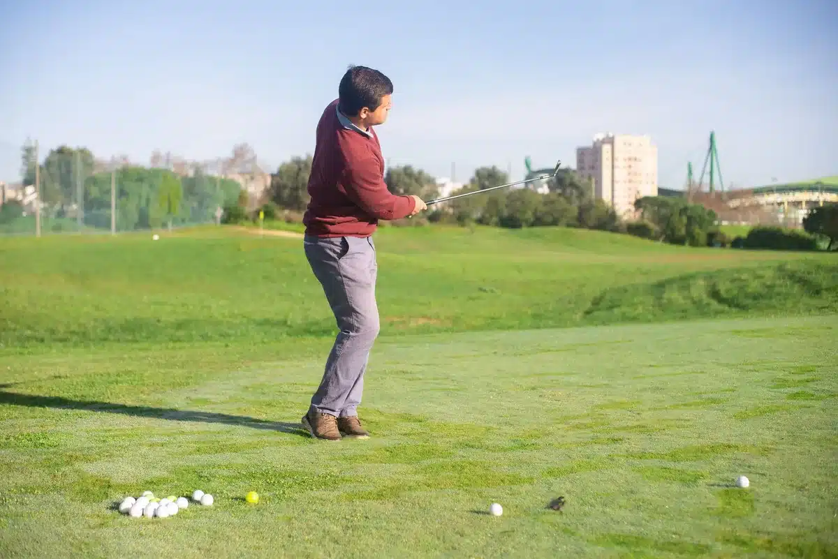 Golfer in mid-swing on a driving range, focused expression, morning light casting long shadows across practice turf, determination evident in posture
