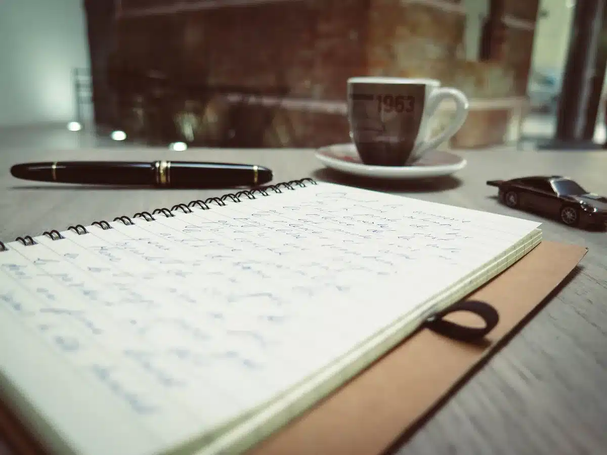 A writing desk with an open notebook, pen poised mid-sentence, soft daylight through a window, a coffee cup nearby, papers scattered with manuscript pages, the scene suggesting creative work in progress