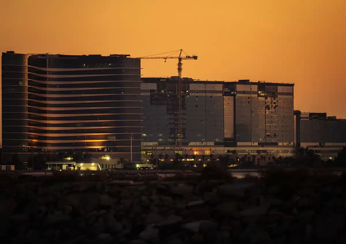 A sprawling data center facility under construction in a high desert landscape, rows of equipment and infrastructure visible, dust and construction activity, early morning light casting long shadows across the site.