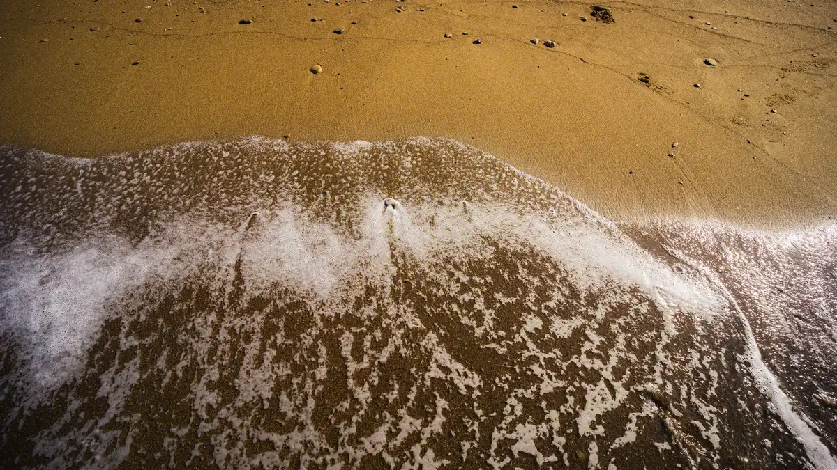 A knife lying on wet sand at the ocean's edge, waves receding, the blade catching sunlight, a stark symbol of threat and danger on an otherwise peaceful beach setting.