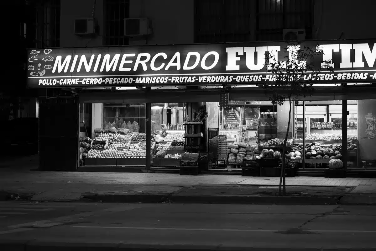 A row of closed storefront signs and darkened windows at a generic supermarket location in an urban shopping district, late afternoon light casting long shadows across the empty parking lot and sealed entrance doors.