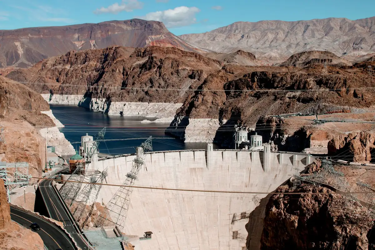 Glen Canyon Dam concrete structure against blue sky, water level far below spillway opening, exposed rock face, minimal water visible in reservoir behind dam