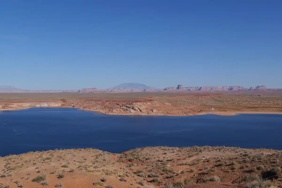 A bathtub-ring watermark on rust-colored rock face showing decades of receding water levels, pale mineral deposits marking the former high-water mark, sparse ripples on the exposed reservoir surface below, dramatic canyon walls rising into clear sky