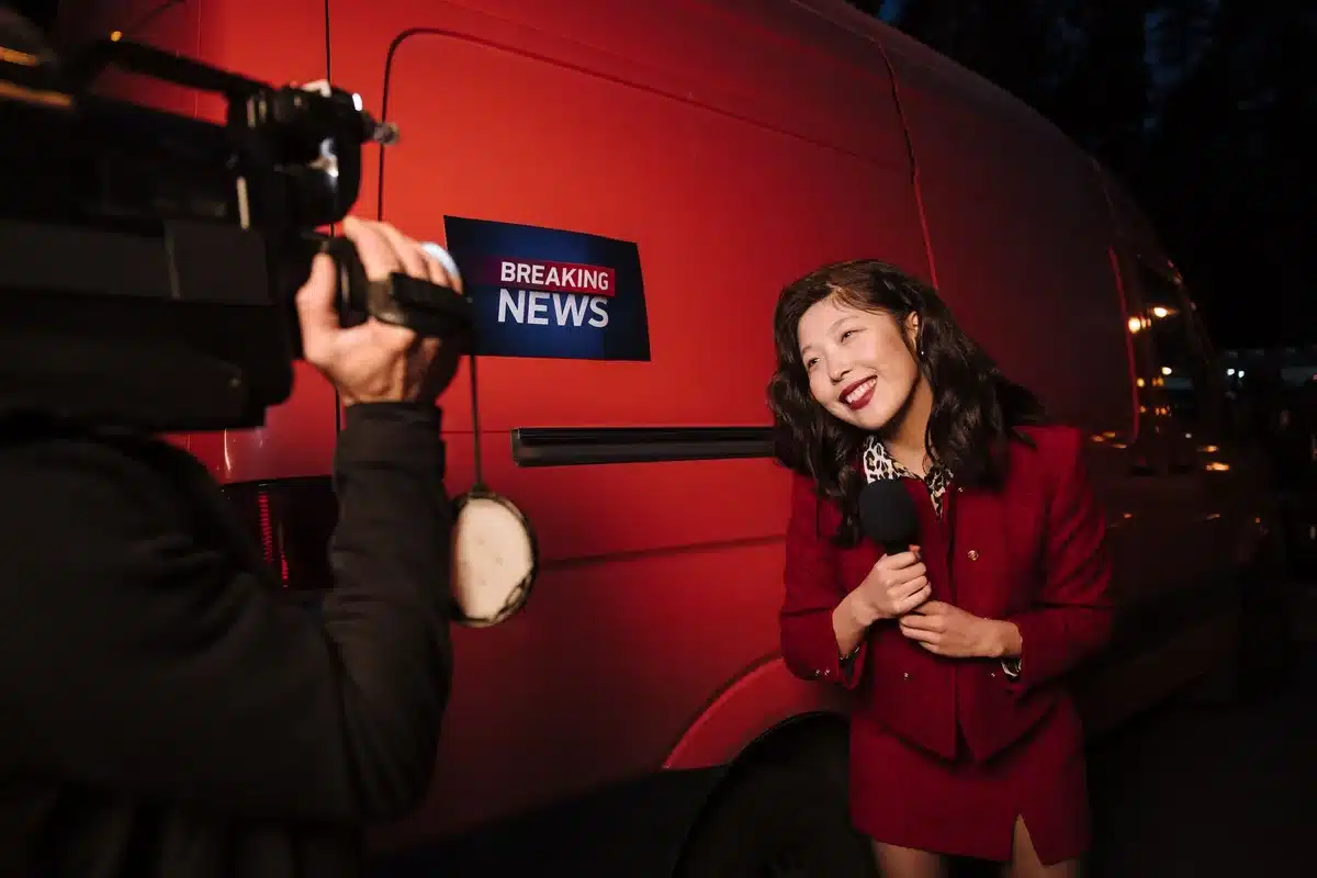 A woman at a podium speaking to reporters, hands gesturing mid-briefing, professional setting with microphones visible