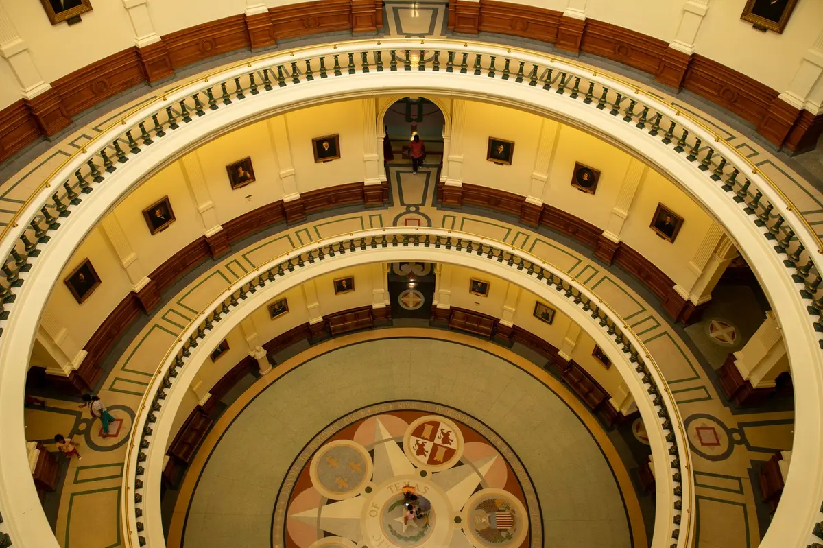 A split-screen showing a podium with a single microphone on the left in dim lighting, and a modern state capitol building on the right in daylight, symbolizing political transition and institutional continuity