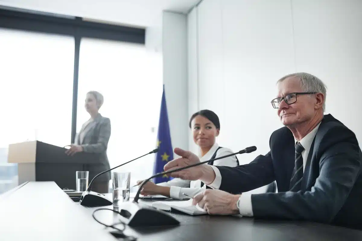 A formal conference room with empty chairs around a long table, harsh overhead lighting casting sharp shadows, documents scattered on the surface, a sense of tense deliberation in an austere government setting