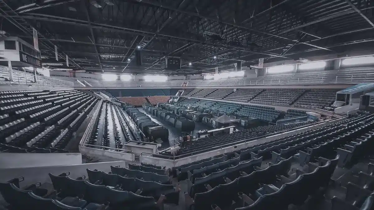 A large public venue interior with empty seats arranged in rows, stage lighting suspended overhead, the scale and grandeur of a major event space, cool architectural tones, a sense of anticipation and scale