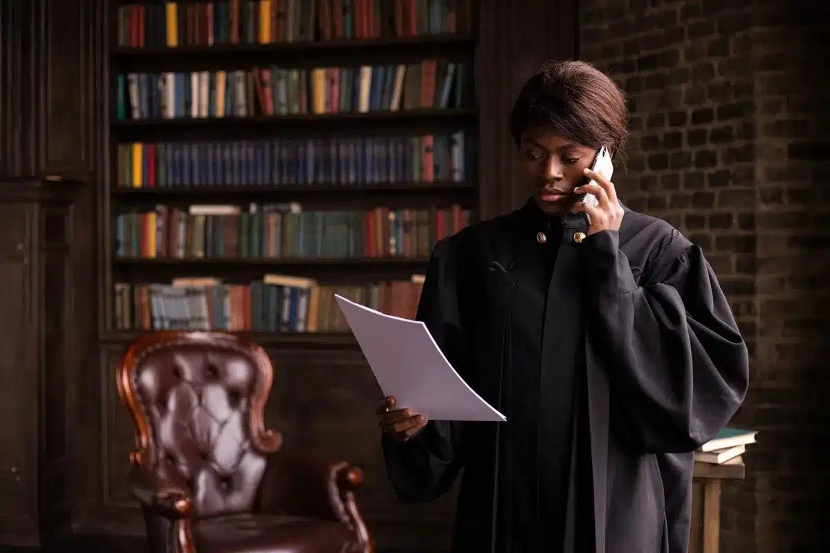 A courtroom interior with empty wooden benches and a judge's bench, harsh overhead lighting casting shadows, a single gavel on the desk, documents scattered on a table, morning light through tall windows