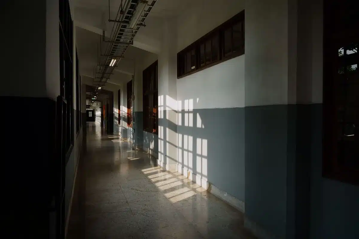 A closed office door with a nameplate, harsh shadows across the hallway, an empty chair visible through a window, fluorescent overhead lighting, a sense of isolation and tension