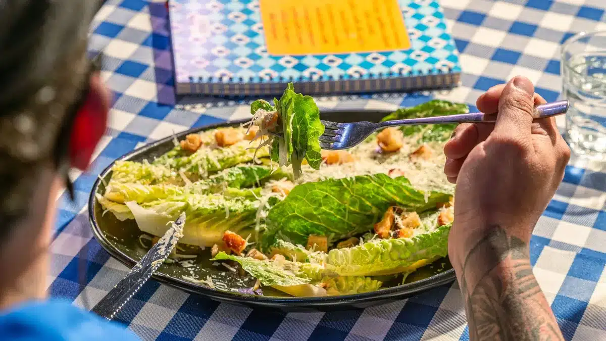 A closeup of creamy Caesar sauce in a white dipping cup, with fresh parmesan shavings and lemon zest visible, set against a blurred fast-food restaurant background.