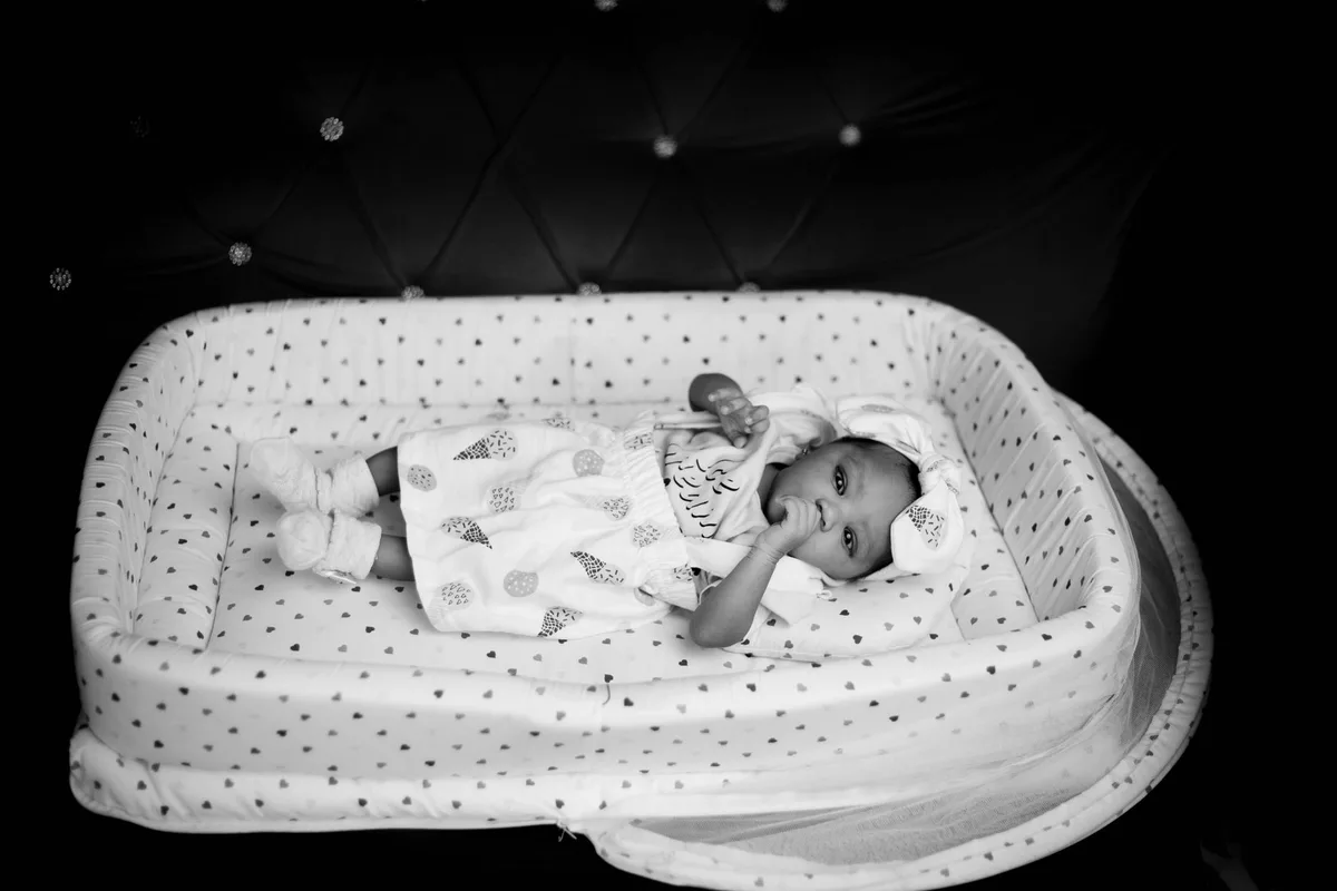A nursery crib beside a stack of policy briefing papers on a desk, morning light streaming through a window, symbolizing the competing demands of parenthood and high-stakes government work.