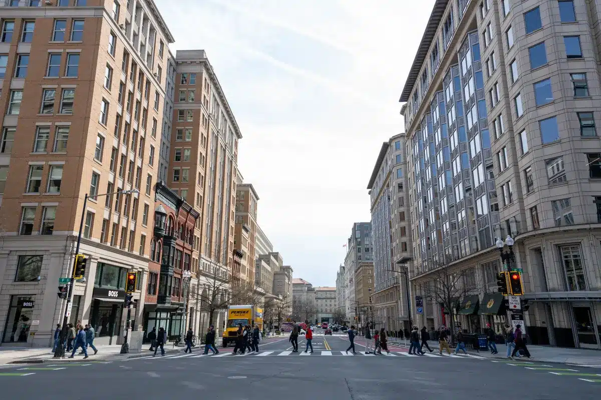 A sprawling street circuit winding through Washington's iconic landmarks, with grandstands packed with spectators lining the National Mall, the Capitol Building visible in the distance, high-speed race cars banking through turns past museums and monuments, bright daylight illuminating the urban racing venue.