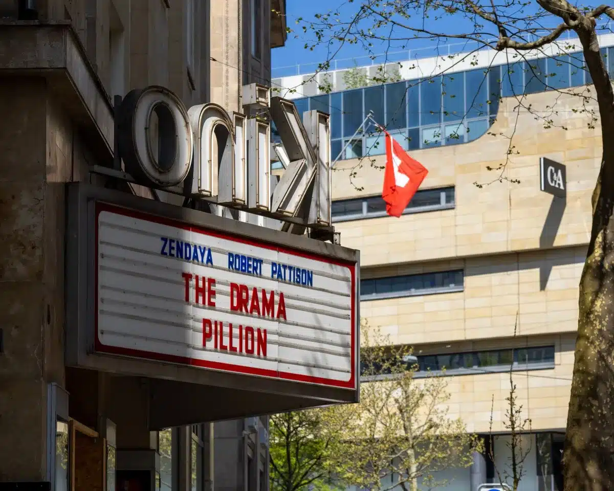 A movie theater marquee displaying a film title in lights, with silhouettes of moviegoers entering, evening ambient light casting long shadows on the pavement.
