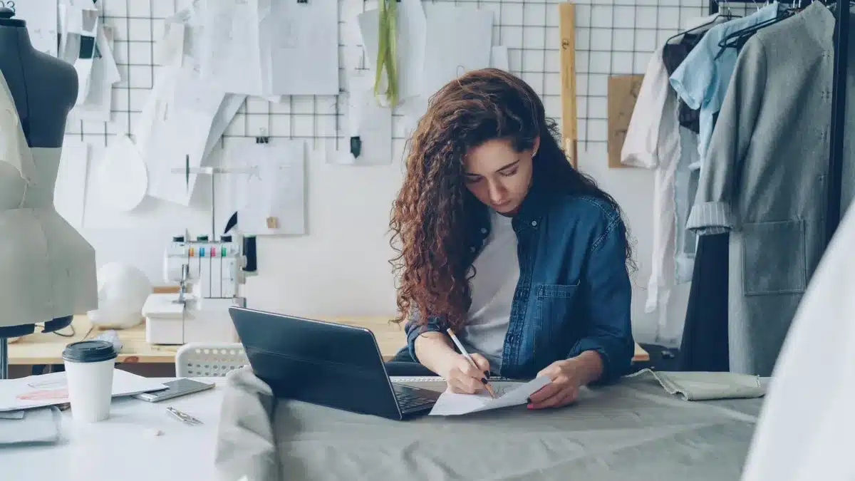 A young designer's sketches and fabric swatches scattered on a design table with measuring tools and a mood board, soft natural light illuminating the creative workspace