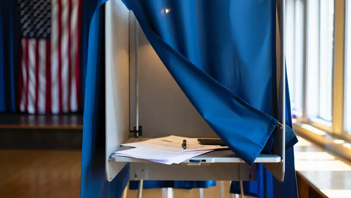 A voting booth with an American flag visible in the background, ballot materials on a small table, morning light streaming through a window, anticipation