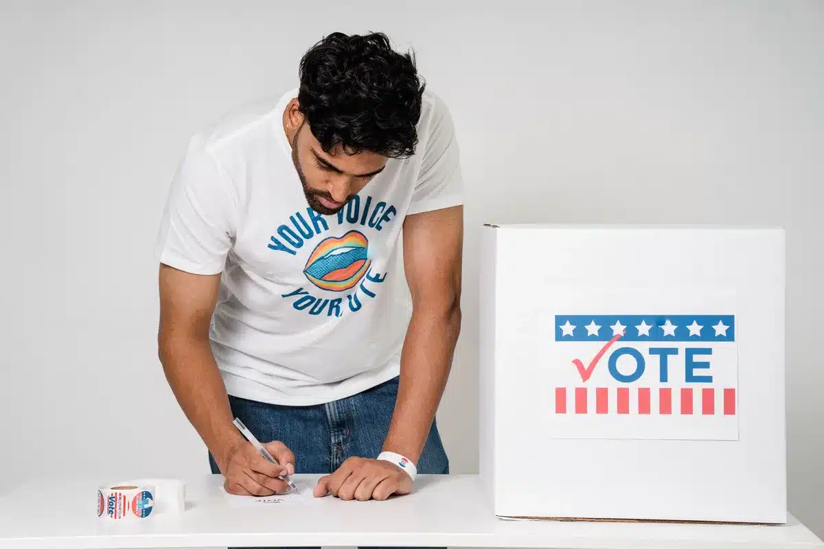An anonymous figure casting a ballot into a voting machine in a quiet polling place, sunlight streaming through windows, a single "I Voted" sticker visible on the edge of the frame