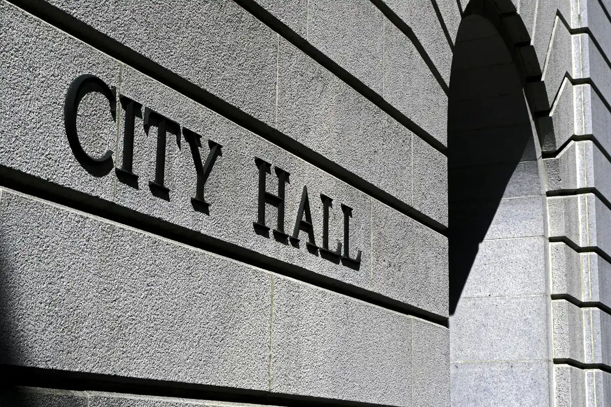 A small-town city hall or municipal building with an American flag at full staff against a clear sky, the flagpole standing firm and solitary, afternoon light casting sharp shadows, quiet civic authority