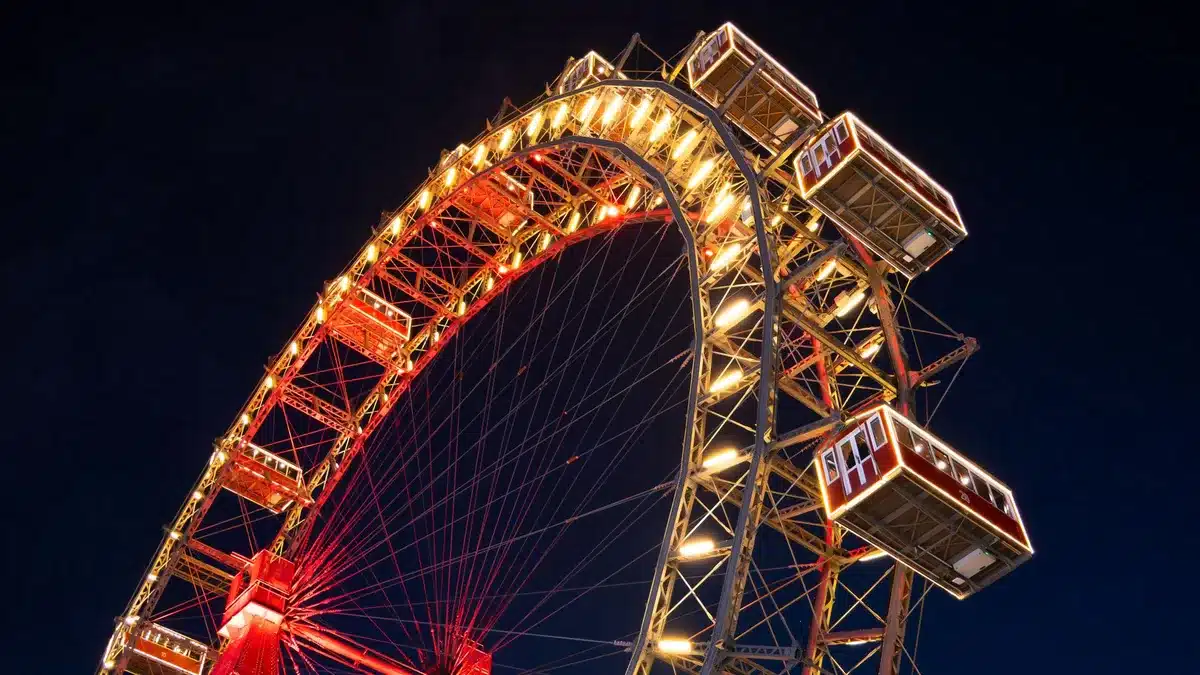 A towering observation wheel with gleaming white structure and enclosed gondolas, silhouetted against a clear blue sky, rising above a sprawling fairground landscape