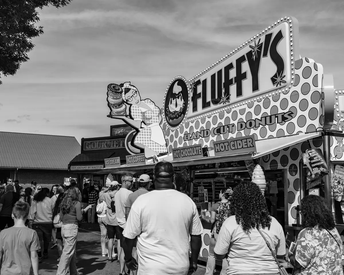 An overhead display of diverse fair foods arranged on wooden boards and paper &mdash; fried items in baskets, colorful drinks in glasses, pastries dusted with sugar, pickled items in jars, all glistening under warm lighting