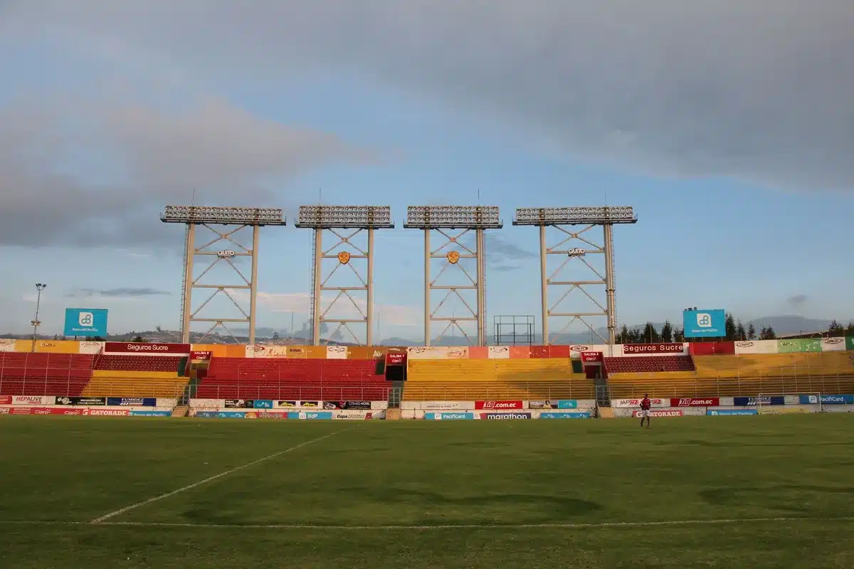 A soccer stadium filled with fans under bright floodlights, a ball suspended mid-air above the pitch during an intense moment of play, goal net visible in sharp focus, crowd emotion visible in the background.