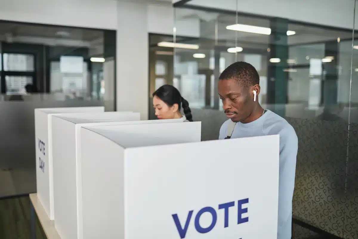 A voting booth interior with a ballot being marked in a polling location, anonymous hands inserting the ballot, soft fluorescent lighting, quiet solemnity