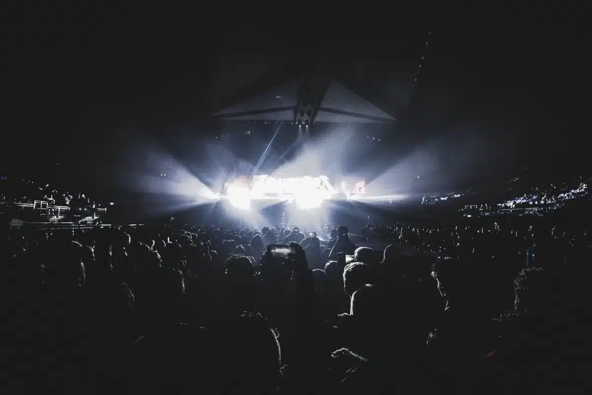 A packed stadium crowd under floodlights, thousands of silhouetted fans with hands raised in unison, faces lost in motion blur, collective energy and elation palpable in the sea of bodies.