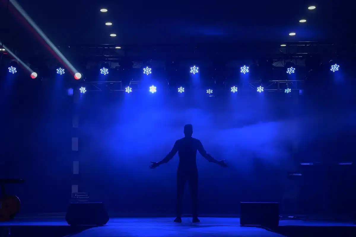 An empty, dramatic concert stage with a single spotlight cutting through darkness, a microphone stand center stage, theatrical fog rolling across the floor, the arena seats dark and waiting.