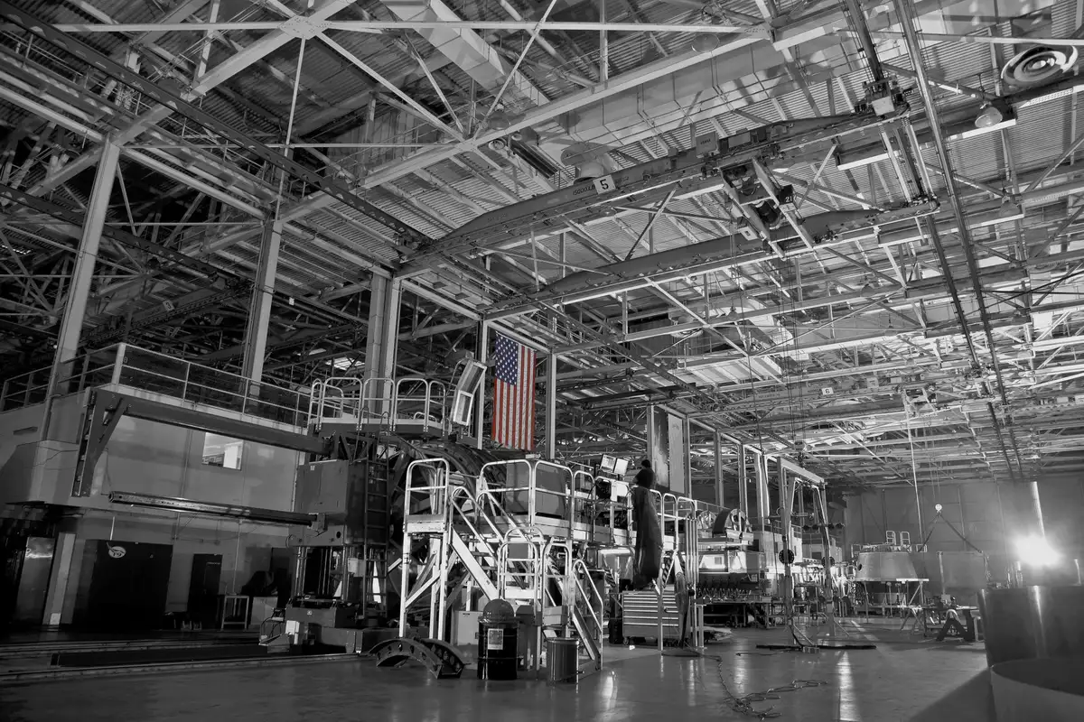 A massive cylindrical spacecraft encased in protective white coverings inside a cavernous clean room facility, technicians in sterile suits performing final inspections, blue and white cables draped across the structure, rows of fluorescent lights overhead
