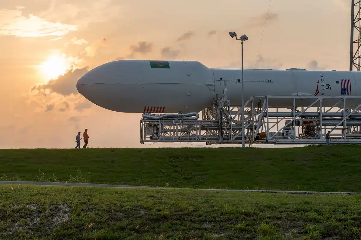 The Falcon Heavy rocket standing vertical on its launch pad at dawn, the three booster cores gleaming white against an orange sunrise, the launch tower visible beside it, a clear Florida sky, wisps of vapor around the base