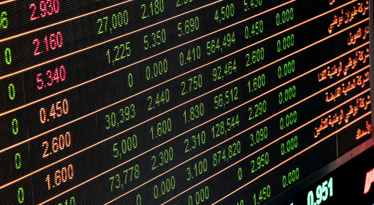 A glowing stock market ticker displaying green numbers and upward-trending candlesticks, with soft blue and green ambient light reflecting off a dark trading desk surface.