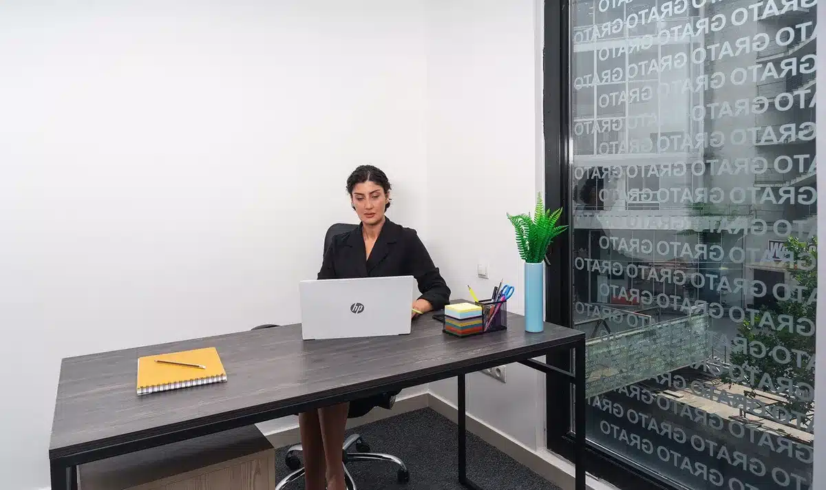 A woman in professional attire sitting at a desk with a laptop, surrounded by papers and printed documents, focused and attentive in a bright office setting