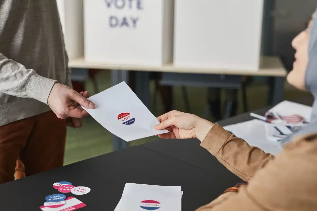 Voter marking ballot at polling place, anonymous hands holding pen over ballot, election booth divider visible, morning light