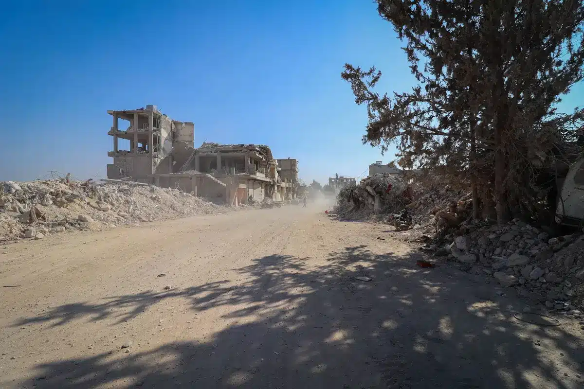 An empty Gaza street with concrete barriers and rubble, Palestinian civilians walking past damaged buildings, Israeli military equipment visible in distance, overcast sky, humanitarian crisis evident