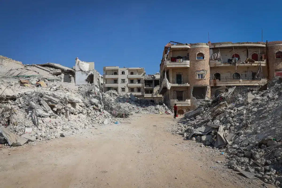 A crowded street in Gaza with residents walking past damaged buildings, sunlight casting long shadows, showing the human cost of prolonged conflict