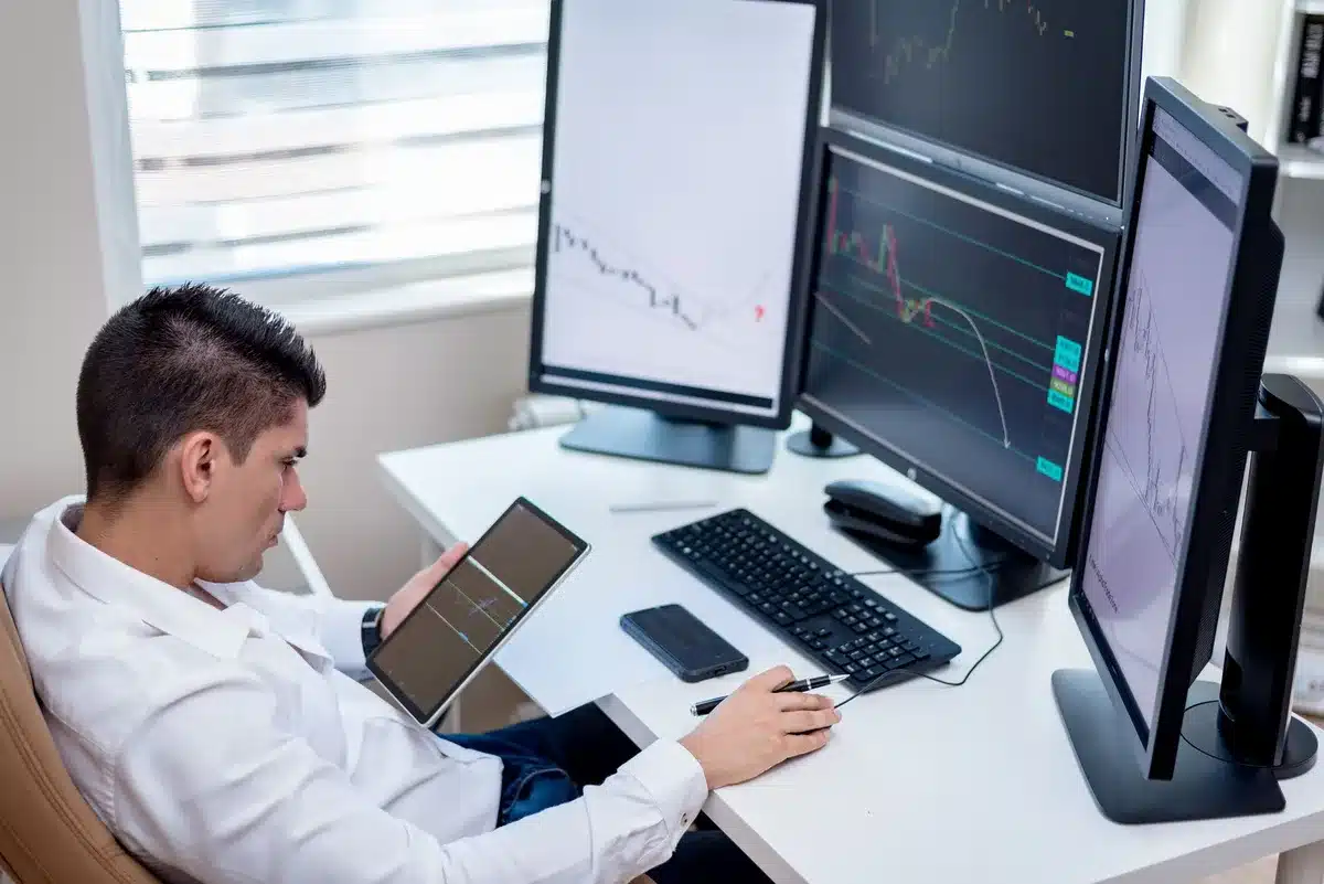 A modern office desk with multiple monitors displaying financial charts and stock tickers, a coffee cup beside a keyboard, warm office lighting casting long shadows