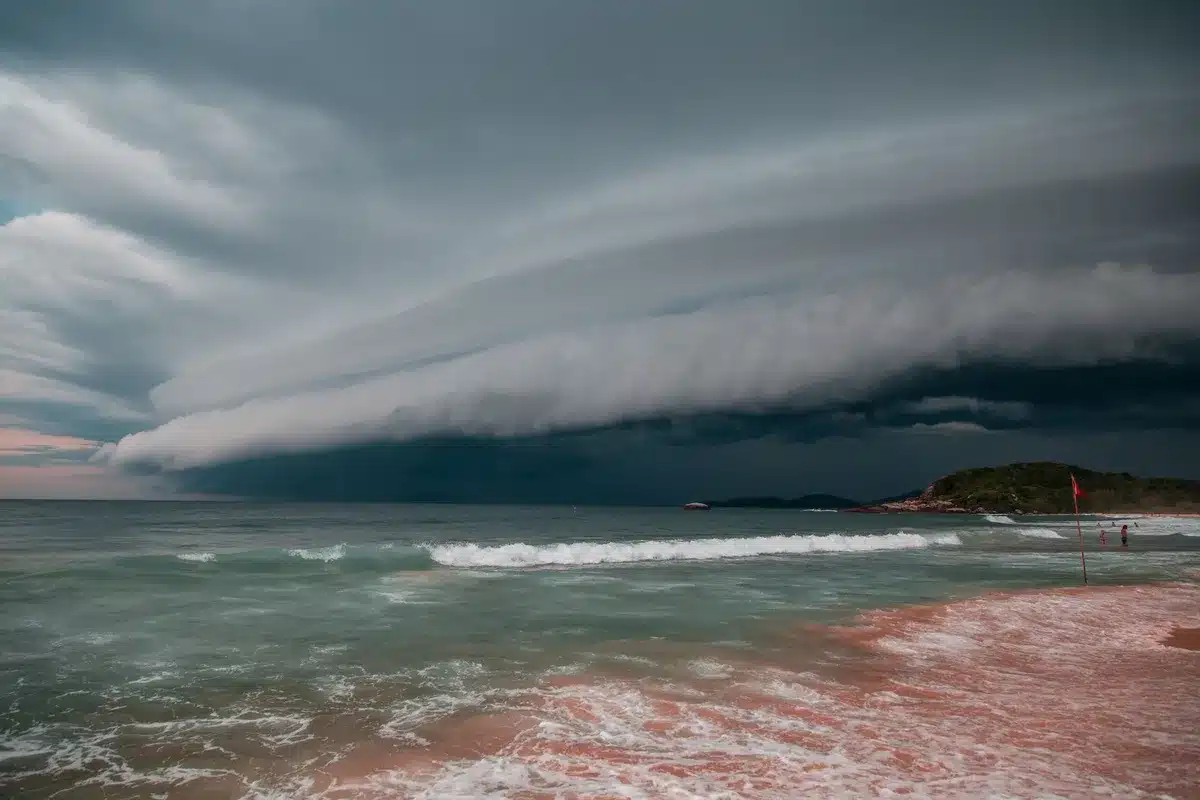 A waterspout visible over ocean water transitioning toward a shoreline with beach structures visible, dark storm clouds above