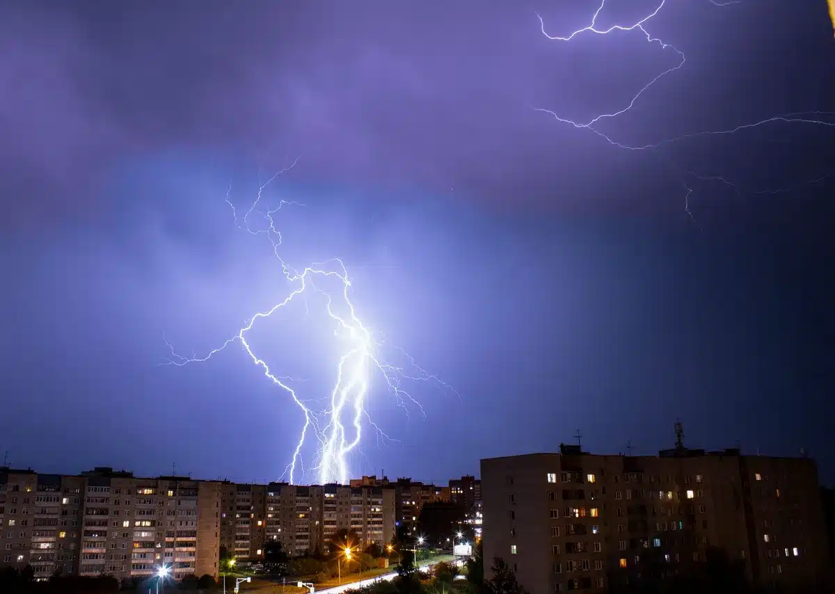 A single lightning bolt striking downward from storm clouds to a silhouetted landscape, intense electric blue-white light against darkness, rain falling in sheets