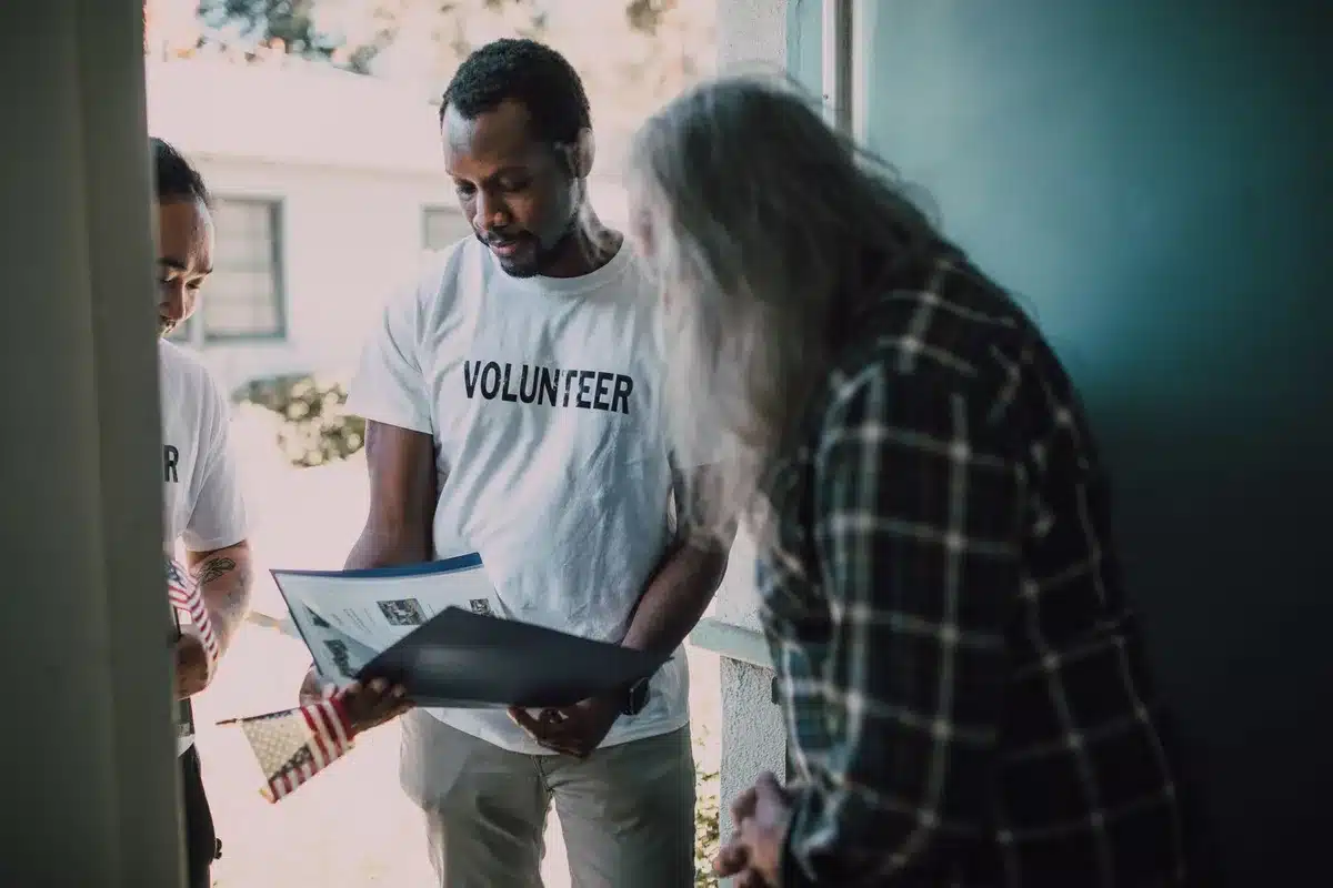 Anonymous volunteers at a community organizing table with voter registration materials and campaign literature spread across a folding surface, engaged in conversation with a resident.
