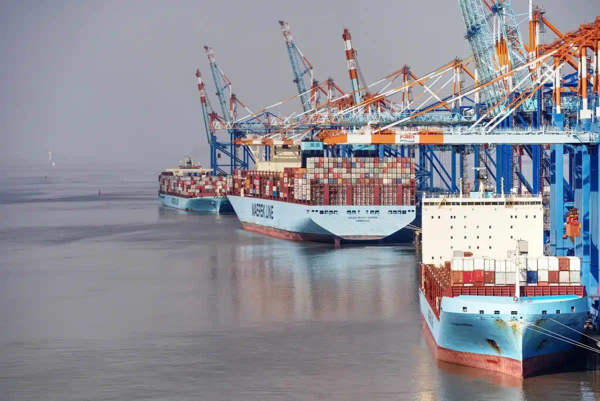 Shipping containers stacked at a port terminal with tanker ships in the background, a hazy skyline, port cranes idle