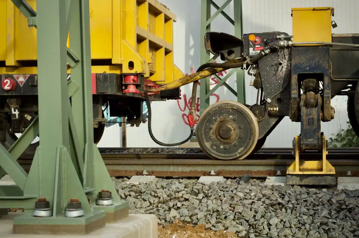 Construction crew working on subway track infrastructure, rail sections stacked nearby, orange safety vests and equipment visible in a tunnel environment
