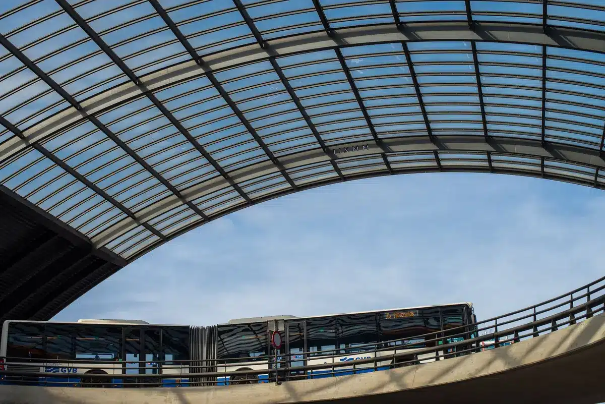 A busy transit station platform with digital signage displaying shuttle bus information, commuters checking phones and schedules, natural station lighting